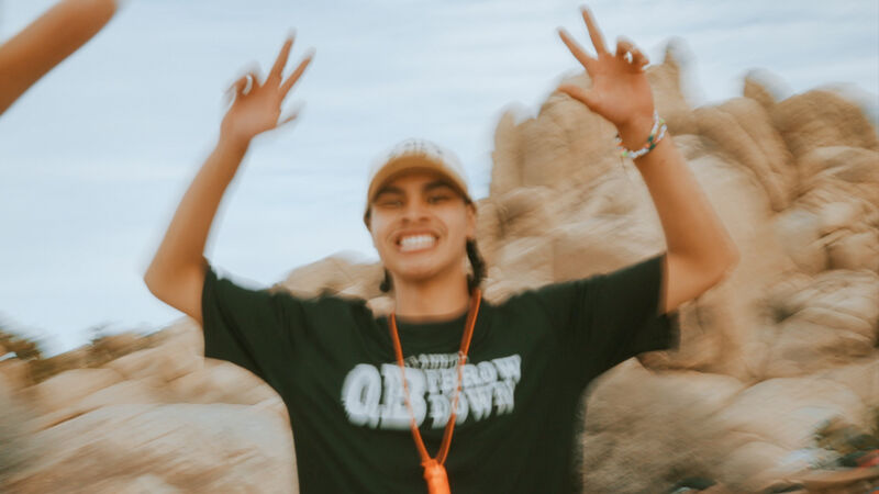 A young man wearing a black t-shirt with the words "OLD TOWN" printed on it, a tan baseball cap, and an orange whistle around his neck is smiling and holding up both hands with two fingers raised on each hand. He appears to be outdoors in a desert-like environment with large rock formations in the background. The image has a slightly blurred effect, suggesting movement or a shallow depth of field.